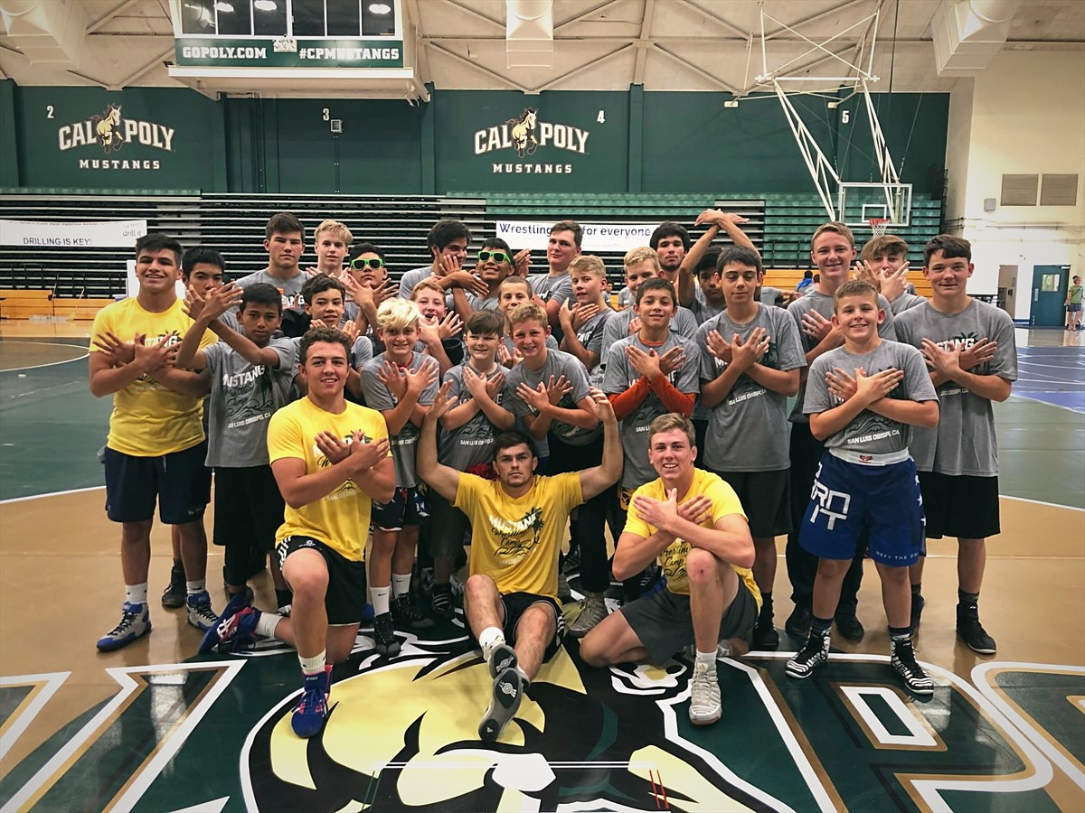 Campers and coaches in camp shirts at center mat in a Cal Poly gym, under the Cal Poly banners